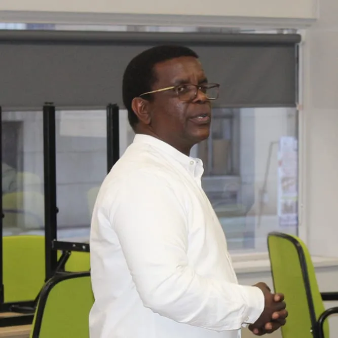 Avo Vision A man in a white shirt and glasses stands in an office with green chairs and large windows, facing slightly away from the camera, possibly preparing for a project management course.
