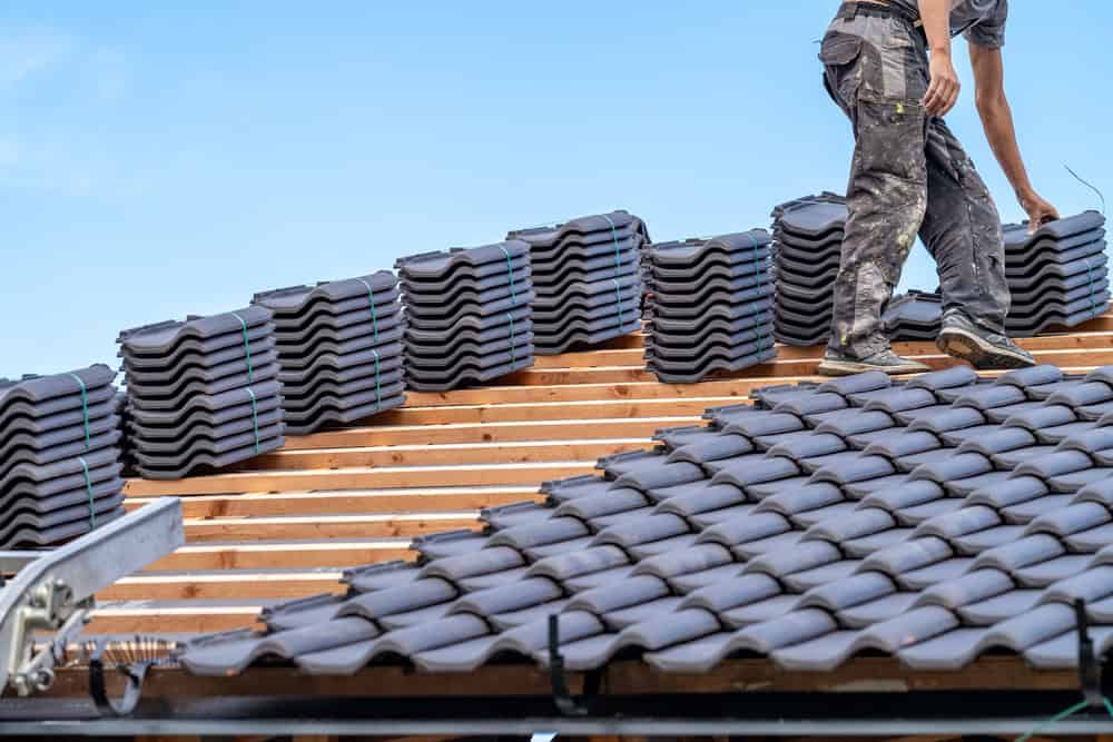 Un artisan couvreur à Valence d’Agen installe des tuiles grises sur un toit, avec des piles de tuiles soigneusement organisées sur la structure en bois à côté de lui.