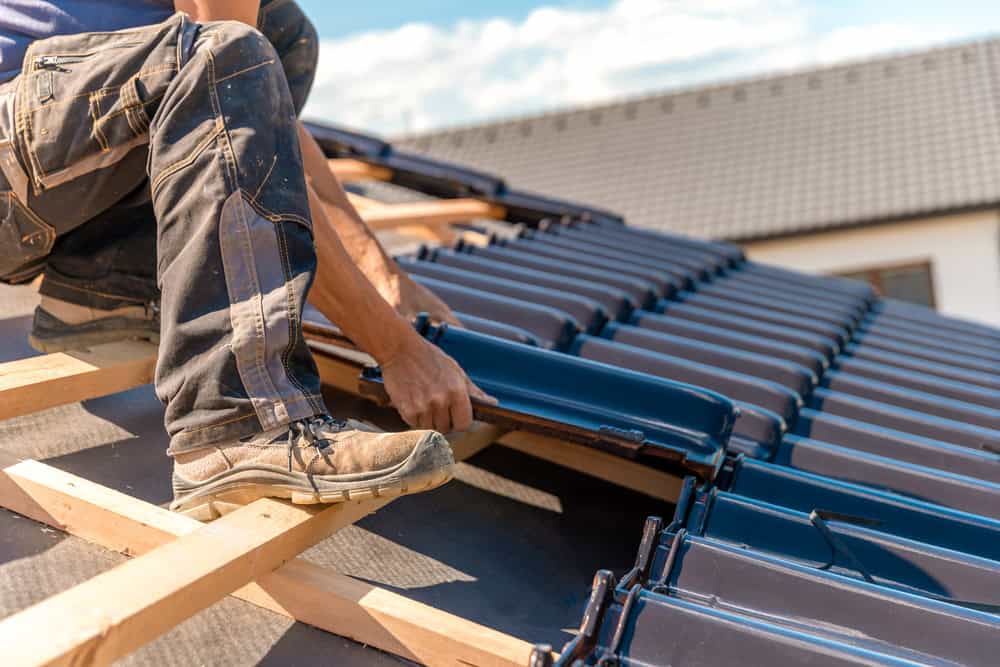 Un couvreur à Montauban, enfilant des bottes de travail et un jean, installe des tuiles bleues sur un toit résidentiel sous un ciel partiellement nuageux.