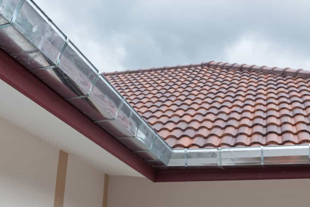 Un toit de maison avec des tuiles en terre cuite rouge et un système de gouttières métalliques, savamment installé par un couvreur zingueur à Caussade, sur fond de ciel nuageux.