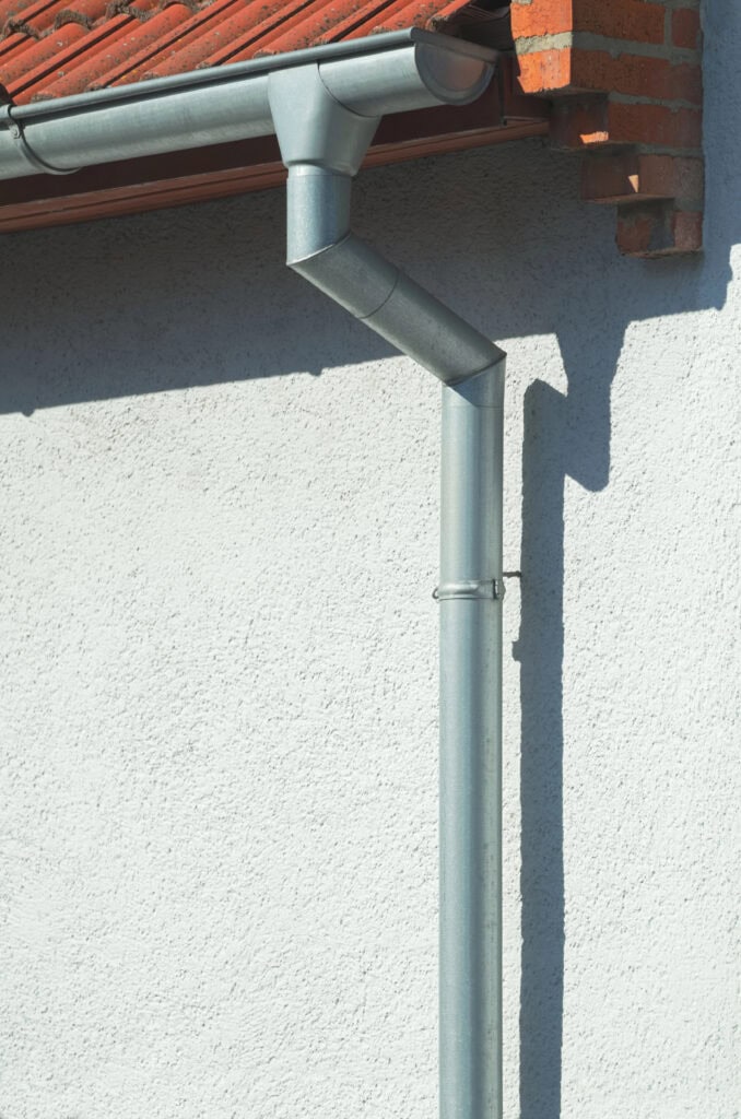 Une descente pluviale en métal gris, savamment installée par un couvreur zingueur à Montbeton, est fixée au mur extérieur en stuc blanc d'un bâtiment au toit de tuiles rouges.