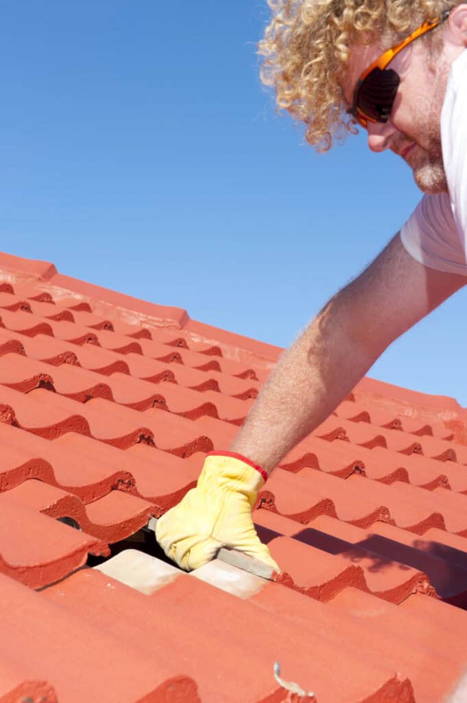Une personne aux cheveux bouclés, portant des lunettes de soleil et des gants jaunes répare ou inspecte un toit de tuiles rouges sous un ciel bleu clair, incarnant le savoir-faire d'une entreprise de couverture à Montauban.