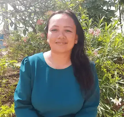 Een vrouw met lang donker haar en een groenblauwe blouse zit buiten voor groene planten en roze bloemen en glimlacht naar de camera op een zonnige dag.