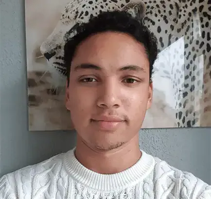 Een jong persoon met kort krullend haar en een witte kabeltrui staat voor een muur met een ingelijste foto van een luipaard op de achtergrond.