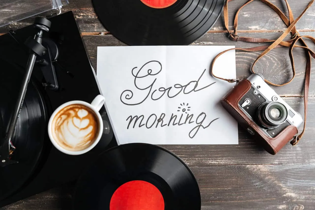 Een handgeschreven goedemorgen op wit papier wordt omringd door een kop koffie met latte art, vinylplaten, een vintage camera met riem en een platenspeler op een houten tafel.