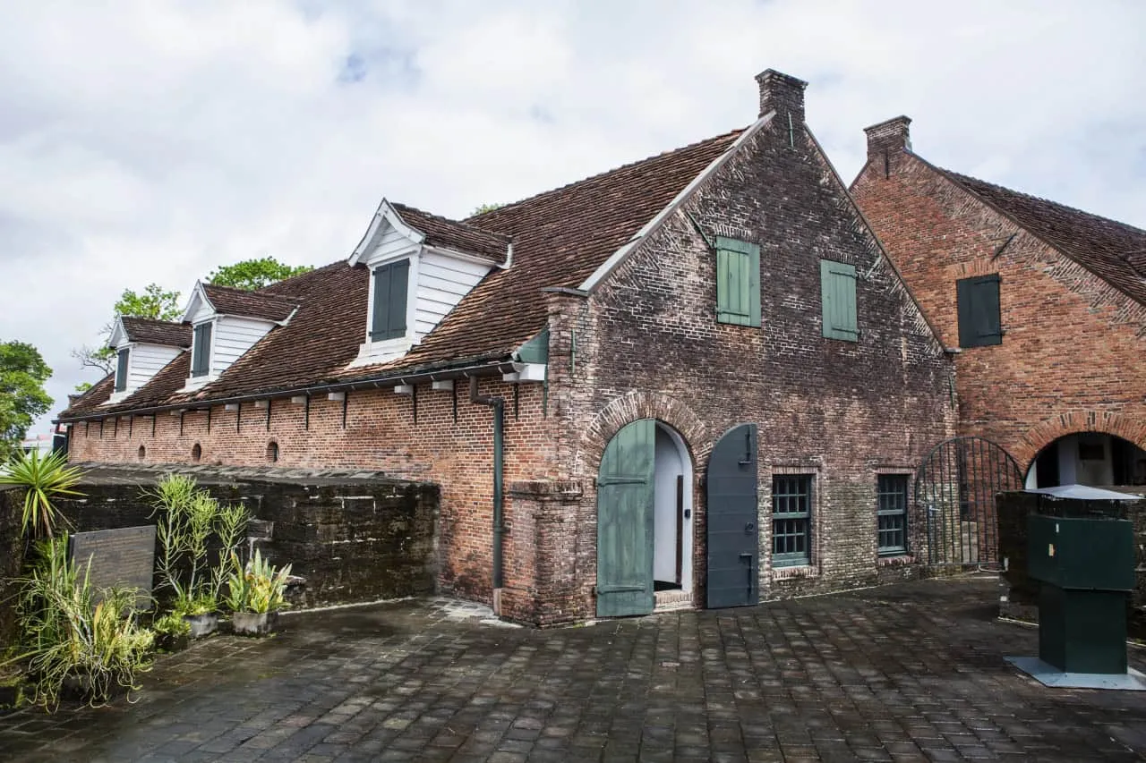 Een historisch bakstenen gebouw in Fort Zeelandia in Suriname, met steile daken bedekt met dakspanen en groene houten luiken, staat op een binnenplaats van natte stenen. In de schaduw van een stenen muur groeien weelderige planten onder een bewolkte hemel.