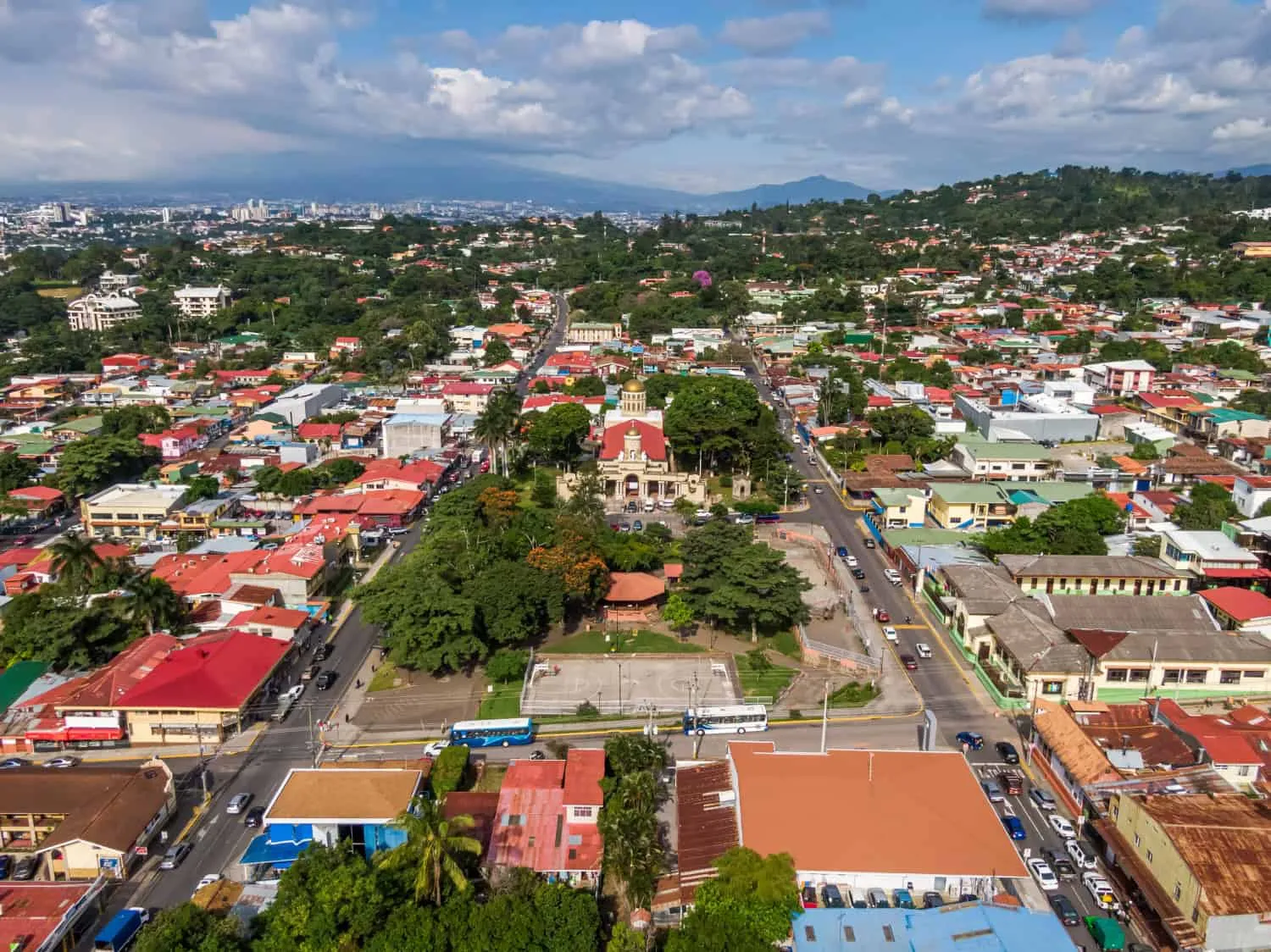Aerial view of Paramaribo city square surrounded by colorful buildings and streets with cars and buses; distant hills and mountains under a partly cloudy sky form the background. Lush trees fill the park in the center of the image.