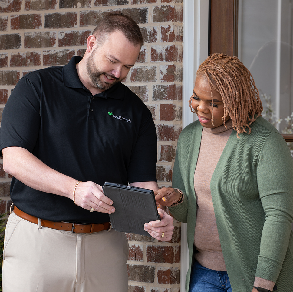 Waynes employee in black polo showing tablet to smiling client at a home's brick entryway