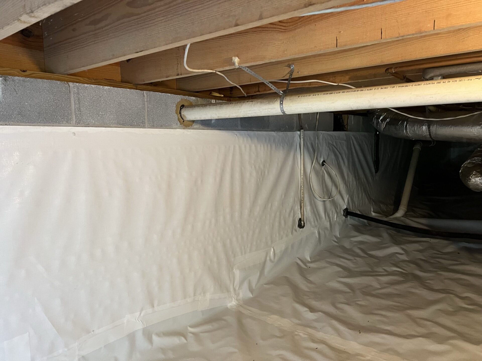 Crawl space with white vapor barrier liner on walls and floor, exposed wooden joists and a suspended PVC pipe