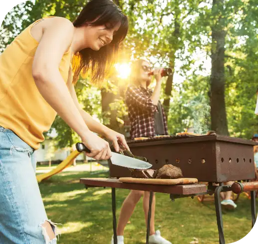 Woman slicing cooked meat on a backyard grill while friends drink and relax in sunny park-like setting