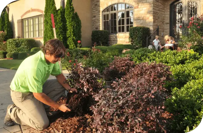 Man kneeling in a front yard scooping soil from a garden bed as a woman and two children sit on the house steps