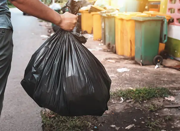 Person carrying a tied black garbage bag past a row of yellow and green outdoor trash bins by the street
