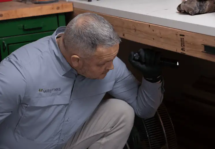 Technician in Waynes uniform crouched under a workbench using a flashlight to inspect a dark area