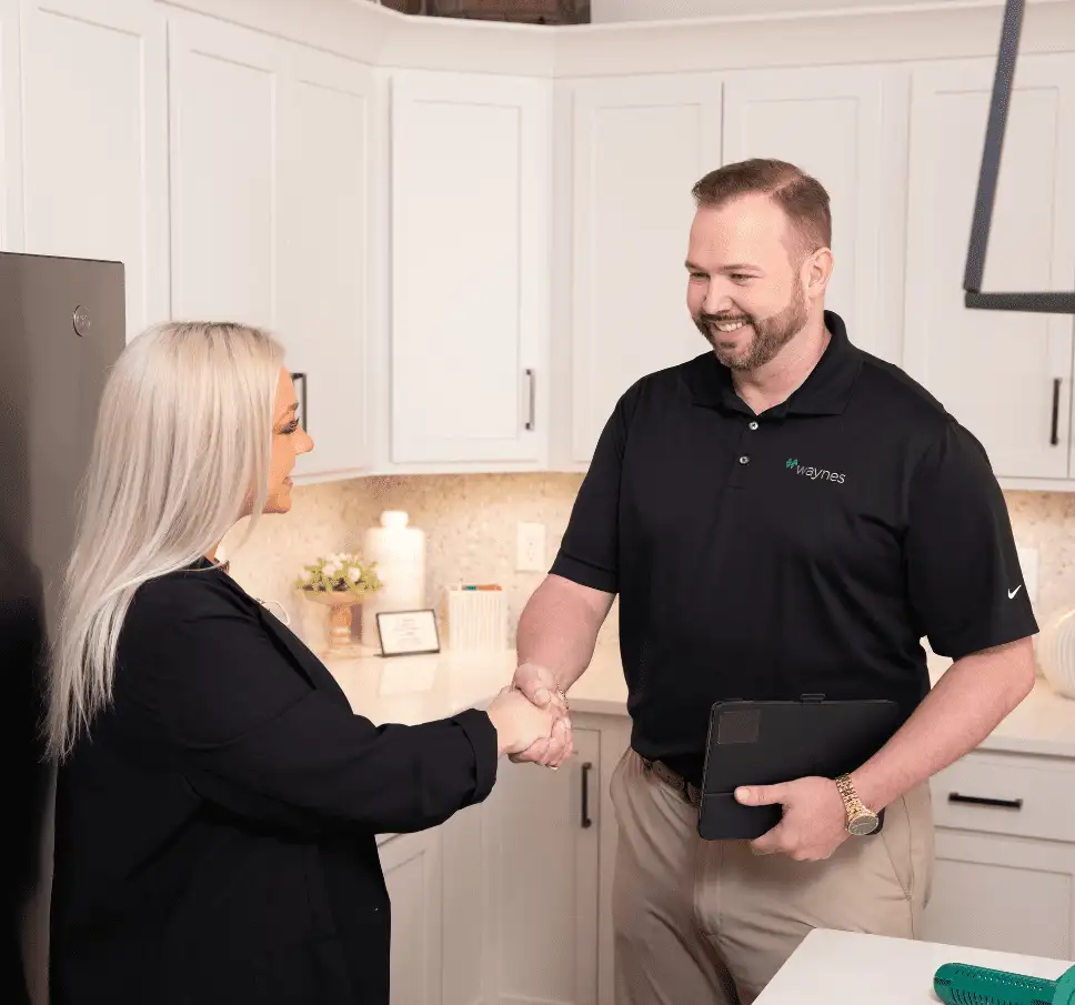 Waynes technician in a black polo shaking hands with a homeowner in a bright kitchen while holding a tablet