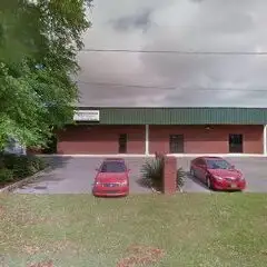 Single-story red-brick commercial building with a green metal roof and two red cars parked on the driveway