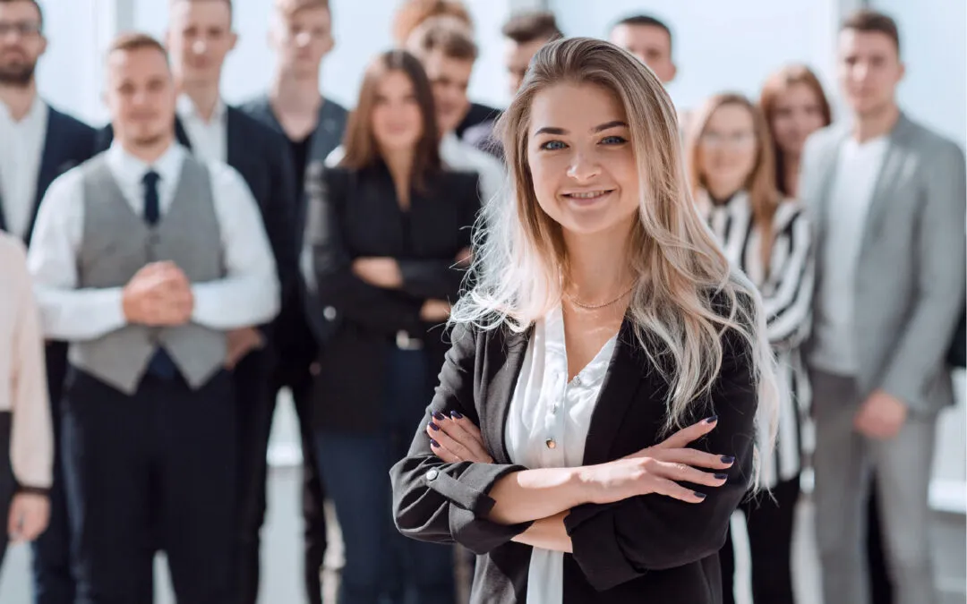 Confident businesswoman standing with arms crossed, supported by a team of colleagues in the background.