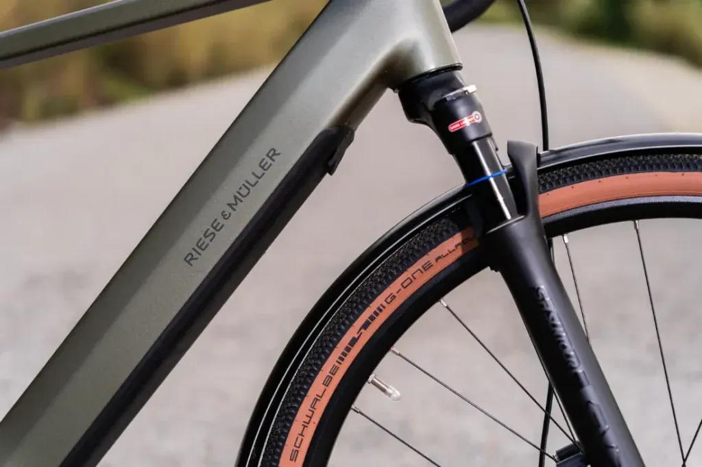 Primer plano de un cuadro de bicicleta Riese & Müller UBN Five con la etiqueta "Riese & Muller", que resalta la ingeniería de precisión de la suspensión delantera y el neumático.