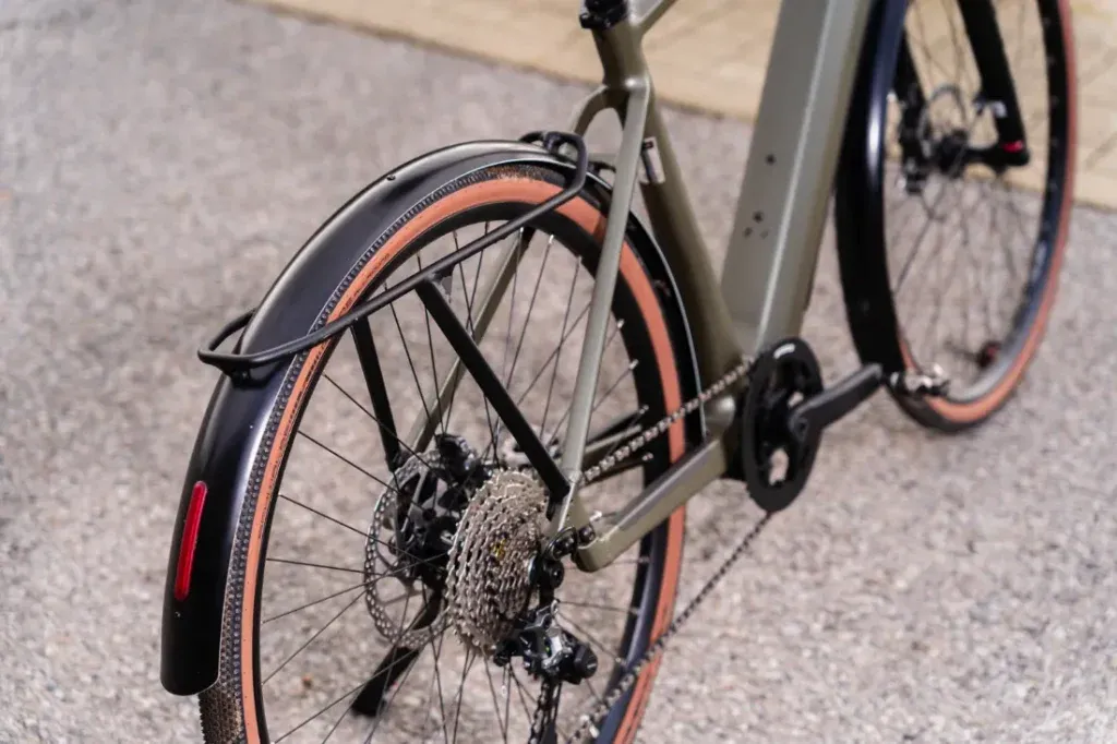 Primer plano de una rueda trasera de bicicleta con un elegante guardabarros urbano, freno de disco y marchas sobre una superficie de grava. Este detalle captura los elementos de diseño innovadores del modelo UBN Five Commute de Riese & Müller.