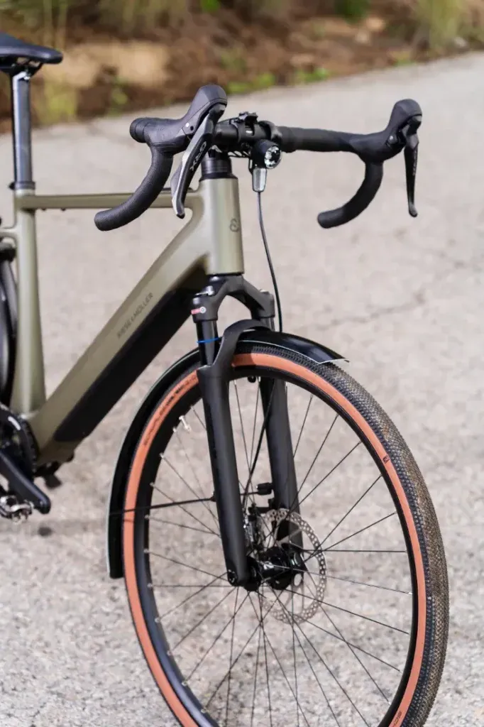 Primer plano de la Riese & Müller UBN Five Commute, que muestra la rueda delantera y el manillar en una carretera asfaltada. El diseño urbano cuenta con un neumático para grava y un freno de disco para un rendimiento mejorado.