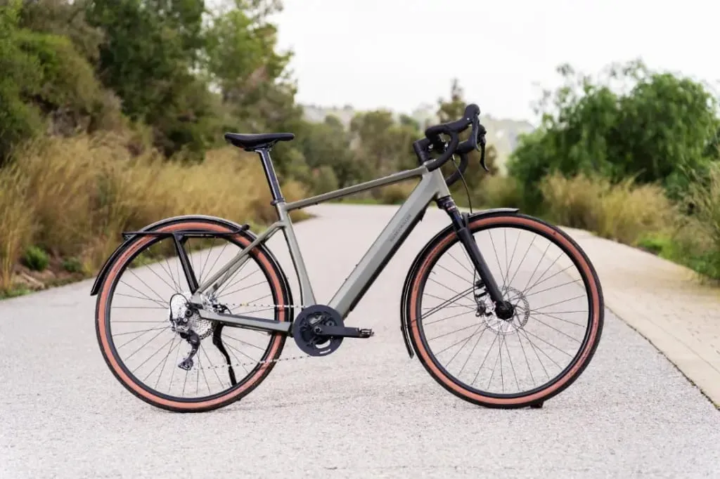Una elegante bicicleta Riese & Müller UBN Five de color gris, con neumáticos con borde rojo, se desliza por una carretera pintoresca y vacía, flanqueada por una exuberante vegetación.