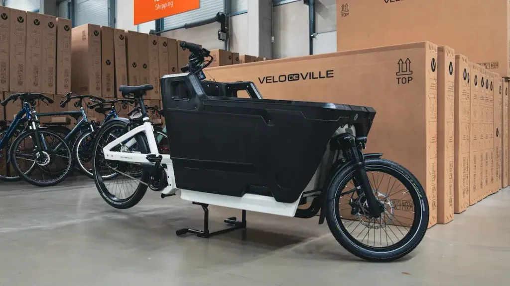 Una bicicleta de carga reposa en el almacén, rodeada de cajas de cartón etiquetadas como "Velo de Ville" junto a otras bicicletas, creando una escena animada que recuerda a los vibrantes mercados de Valencia.