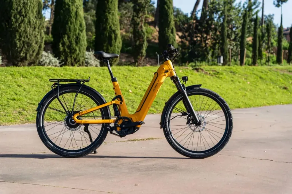 La elegante FEB 990 de Velo de Ville, una bicicleta eléctrica de color amarillo vibrante, está estacionada en un camino pavimentado, rodeada de exuberante césped verde y árboles imponentes.