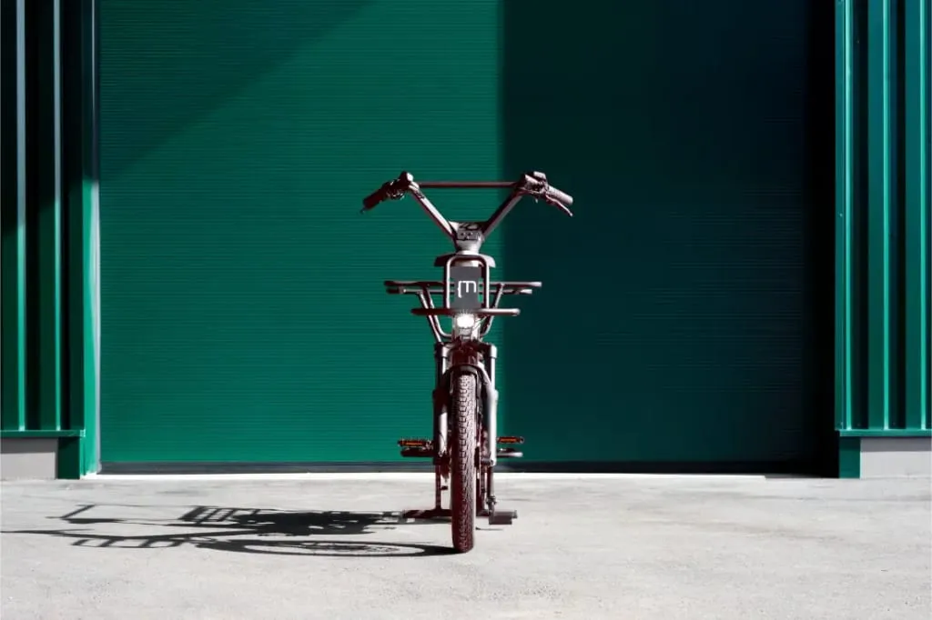 Una bicicleta roja Moustache Lundi 20 Cargo está estacionada mirando hacia adelante contra una puerta de garaje verde bajo la brillante luz del sol.