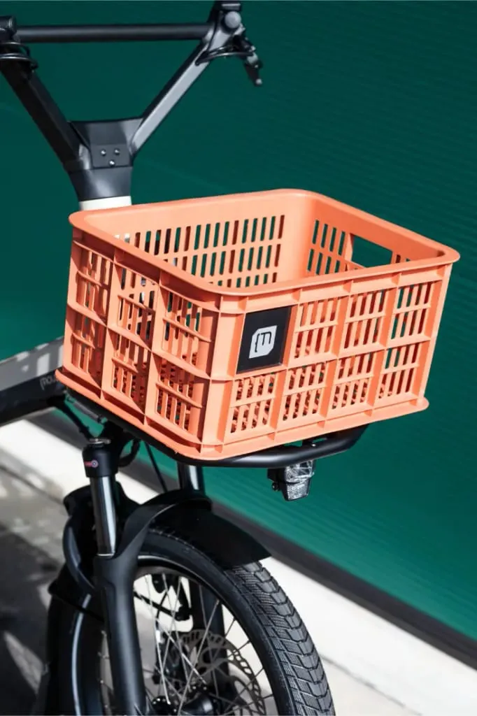 Una caja naranja está unida a la parte delantera de una bicicleta Moustache Lundi 20 Cargo negra, colocada contra una pared verde.