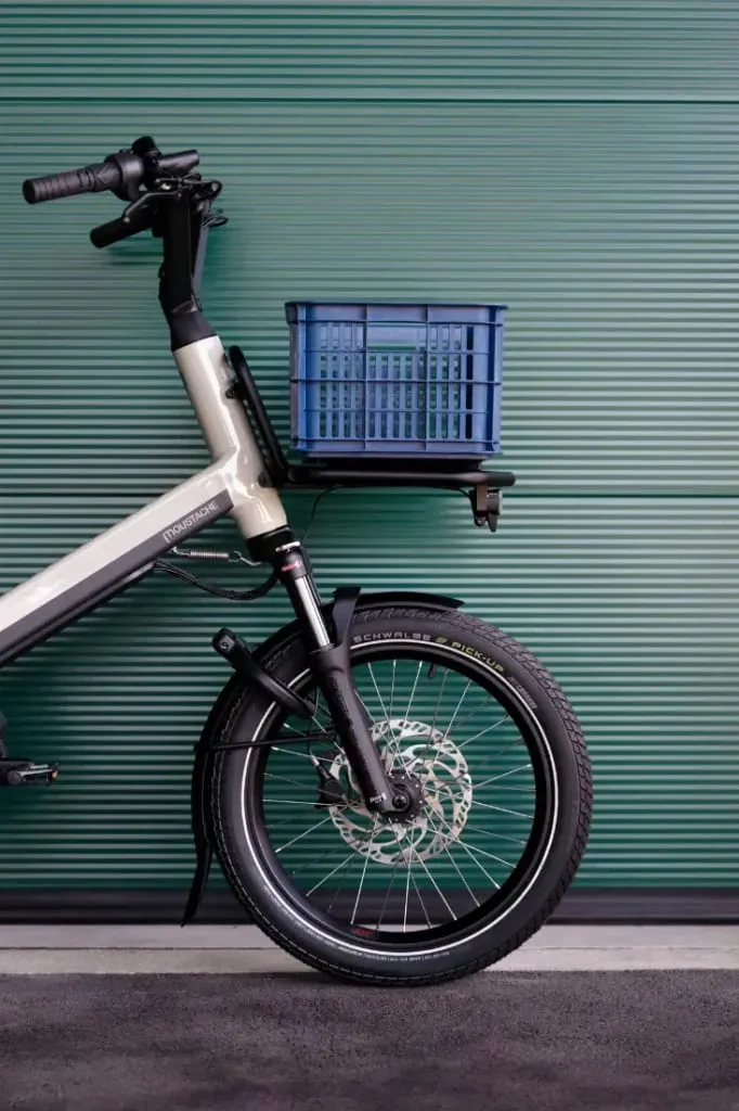 Vista frontal de una bicicleta de carga Moustache Lundi 20 con un cajón azul colocado encima de la rueda delantera, sobre un fondo verde.