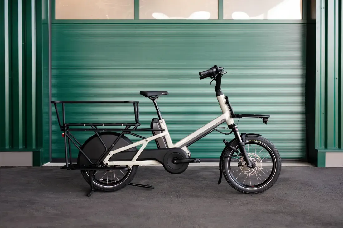 Una moderna bicicleta eléctrica Moustache Lundi 20 Cargo con un portaequipaje trasero resistente, estacionada frente a una puerta de garaje verde.