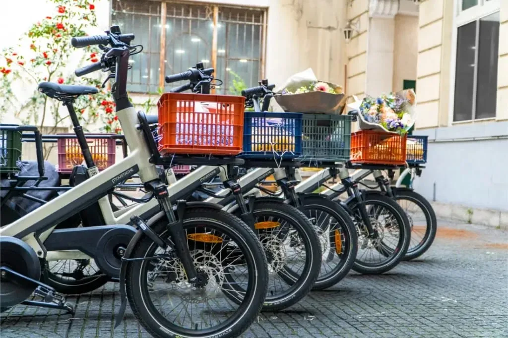 Cuatro bicicletas, incluida una Moustache Lundi 20 Cargo con una cesta delantera llena de flores, están alineadas en una calle adoquinada cerca de un edificio.