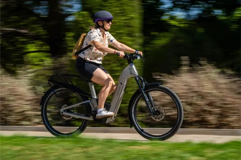 Con casco y mochila, una persona conduce rápidamente una bicicleta eléctrica Riese & Müller Homage5 por un sendero, con vegetación de fondo.