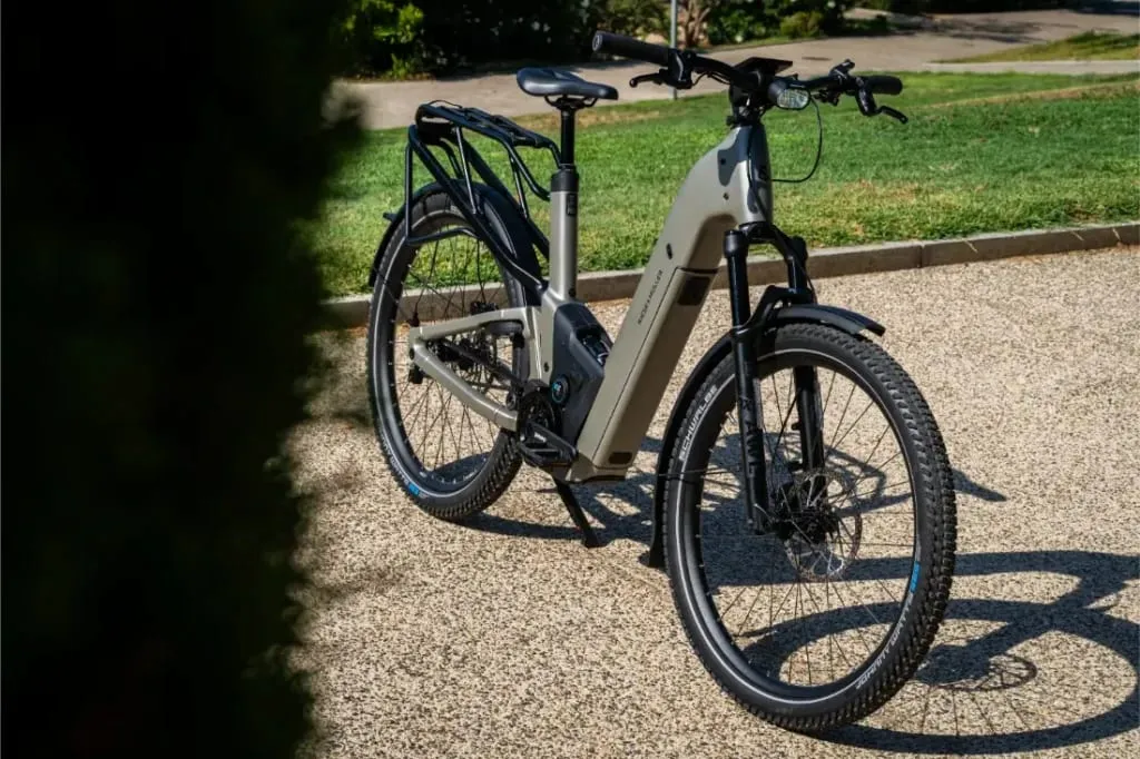 Una moderna bicicleta eléctrica Riese & Müller Homage5 GT con cuadro de paso bajo está aparcada en un camino de grava, rodeada de hierba y árboles al fondo.