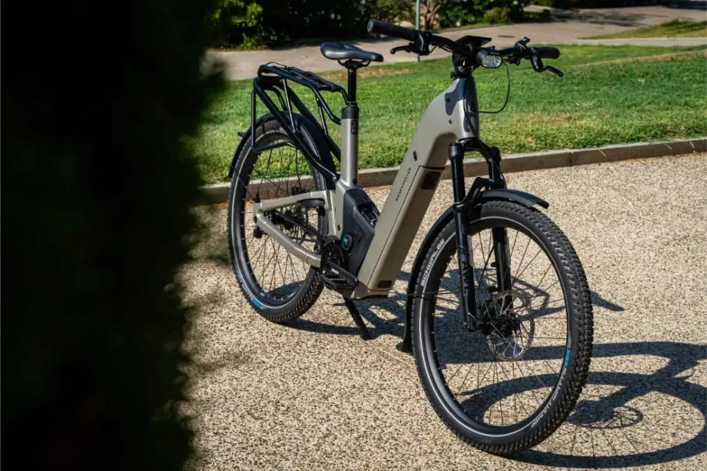 Una moderna bicicleta eléctrica de piñón Riese & Müller Homage5 GT con cuadro de paso bajo está aparcada en un camino de grava, rodeada de hierba y árboles al fondo.