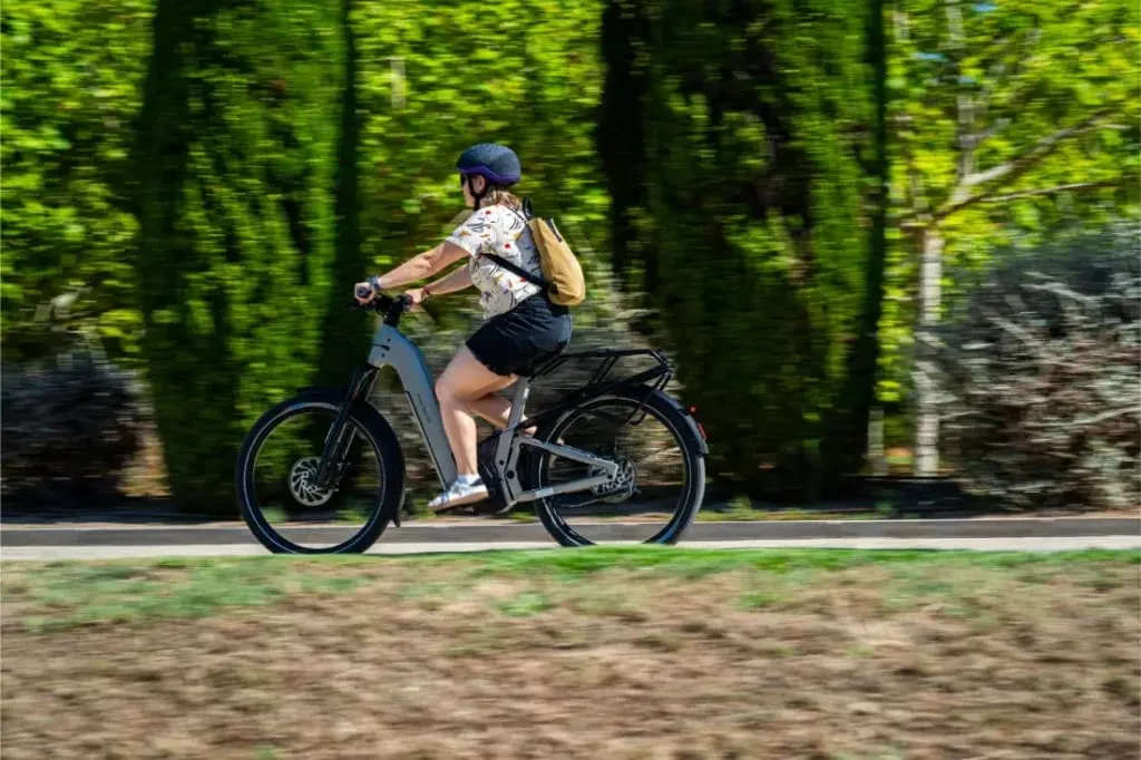 Con casco, una persona monta en una Riese & Müller Homage5 con paso bajo por un sendero bordeado de árboles bajo el sol.