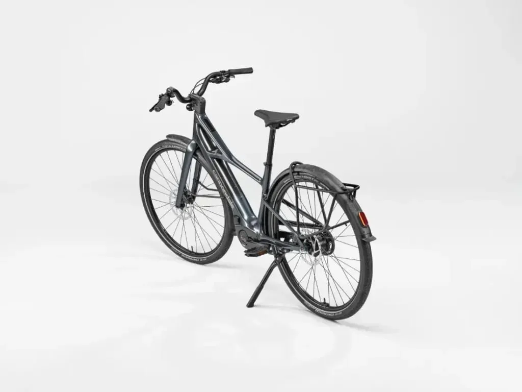 Una bicicleta eléctrica Cannondale FlyingV gris con guardabarros y pata de cabra, vista desde atrás a la izquierda sobre fondo blanco.