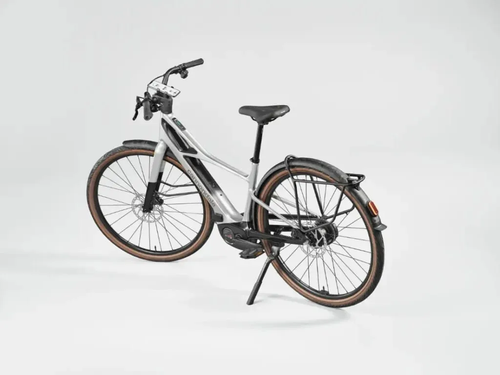 Bicicleta eléctrica Cannondale FlyingV plateada con sillín y neumáticos negros, apoyada en su caballete sobre un fondo blanco.