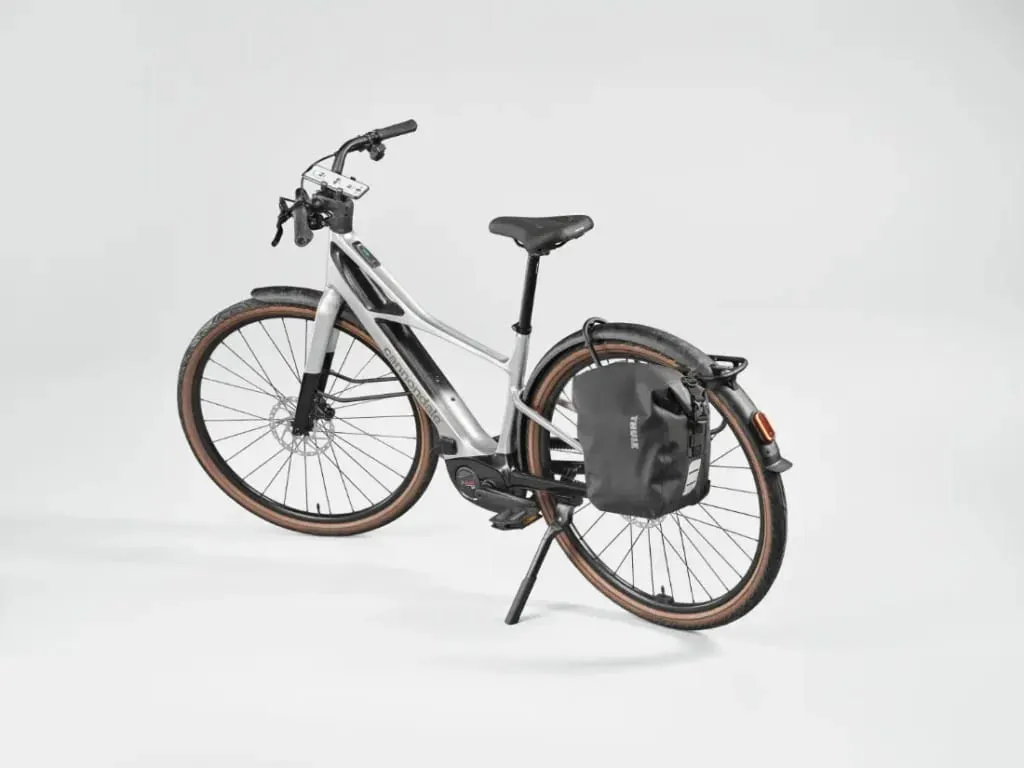 Elegante bicicleta eléctrica Cannondale FlyingV plateada con neumáticos marrones, portabultos trasero y una bolsa negra, sobre fondo blanco.