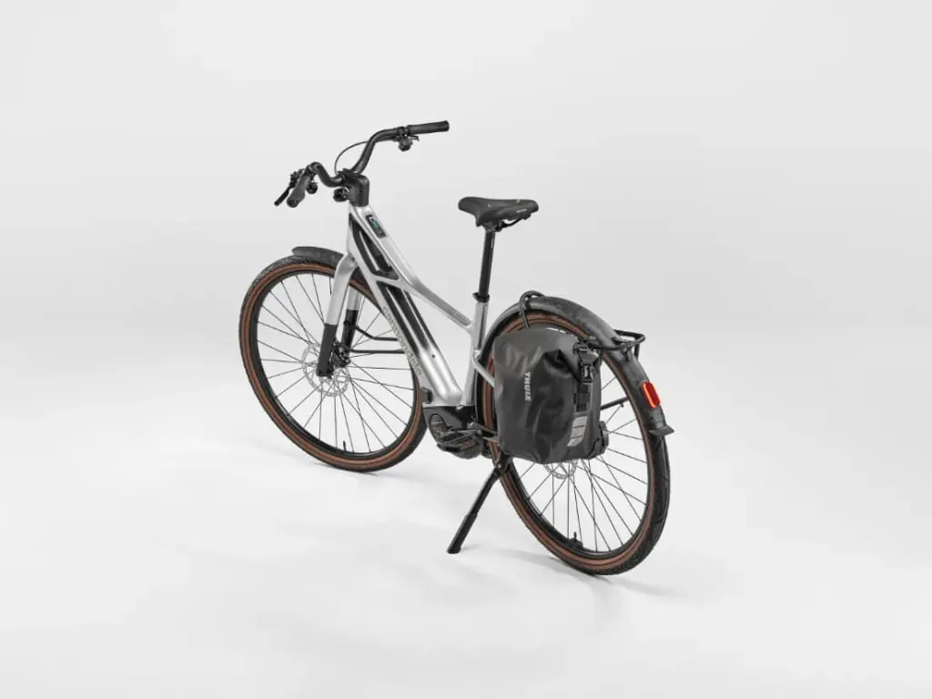 Bicicleta eléctrica Cannondale FlyingV plateada con neumáticos marrones, un portaequipajes trasero y una bolsa negra tipo alforja, mostrados sobre un fondo blanco.