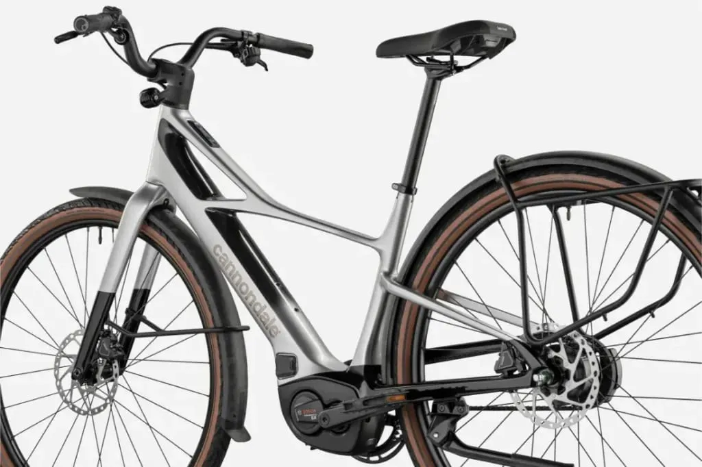 Bicicleta eléctrica Cannondale FlyingV plateada con neumáticos gruesos, portabultos trasero y una batería visible montada en el cuadro, vista lateral.