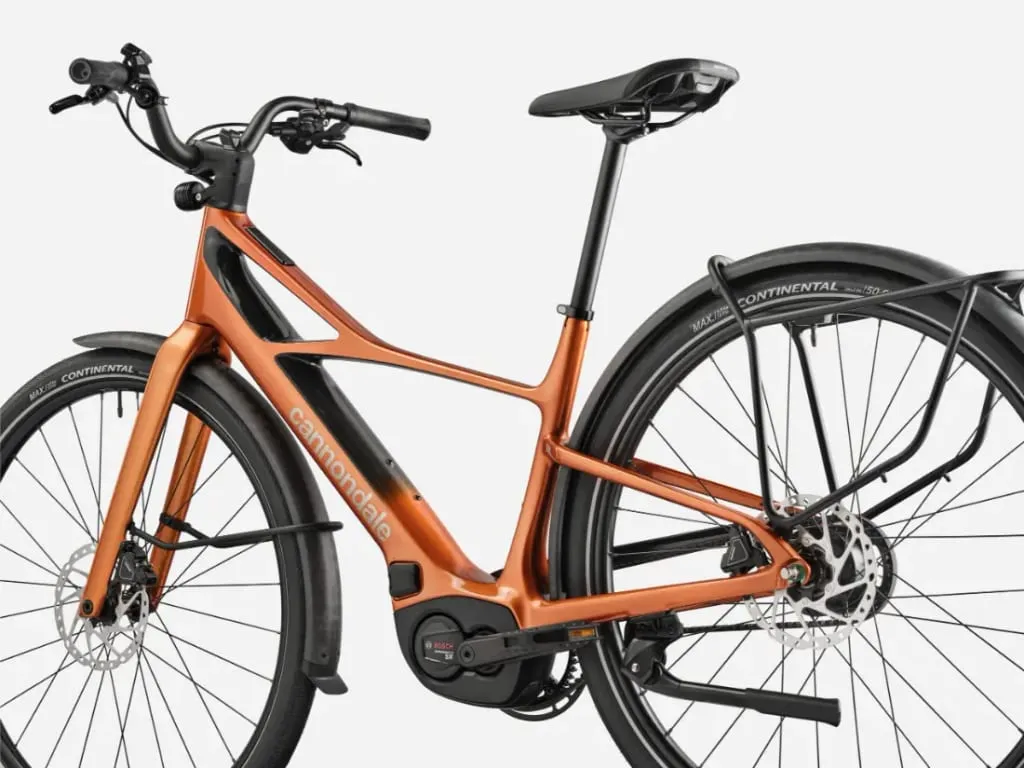 Una bicicleta eléctrica Cannondale FlyingV de color naranja metalizado con neumáticos negros, guardabarros y un portabultos trasero, mostrada de lado.