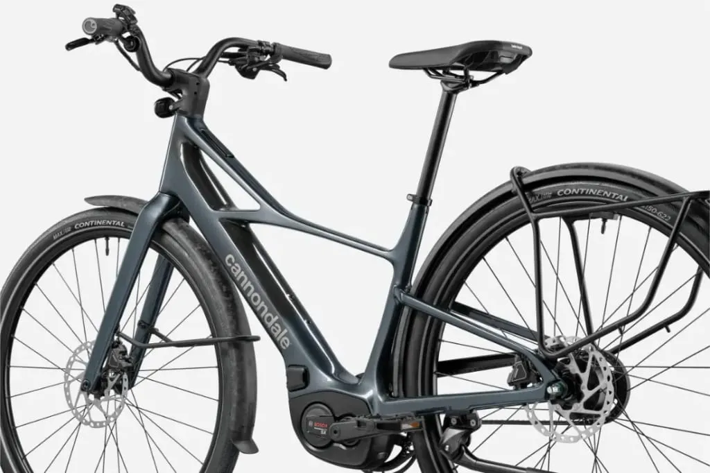 Una elegante bicicleta eléctrica Cannondale FlyingV gris con neumáticos negros, portabultos trasero y motor visible sobre fondo blanco.