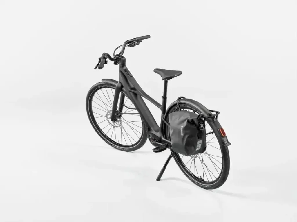 Una bicicleta eléctrica negra mate con una bolsa trasera, vista desde atrás a la izquierda sobre un fondo blanco.