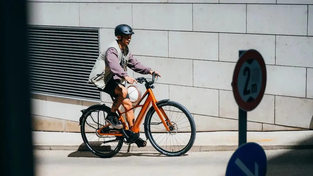 Una persona con casco conduce una bicicleta Cannondale FlyingV naranja por una calle de la ciudad, cerca de señales de tráfico y de un muro de baldosas.