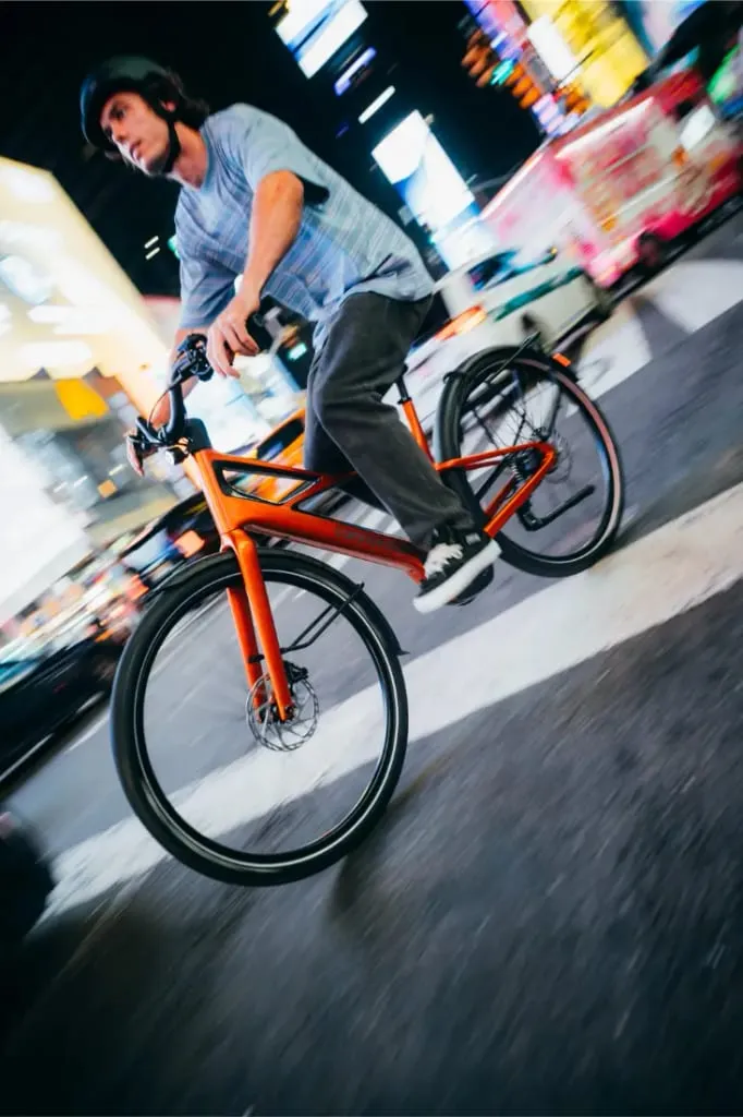 Una persona con casco monta en una bicicleta Cannondale FlyingV naranja por una calle de la ciudad de noche.