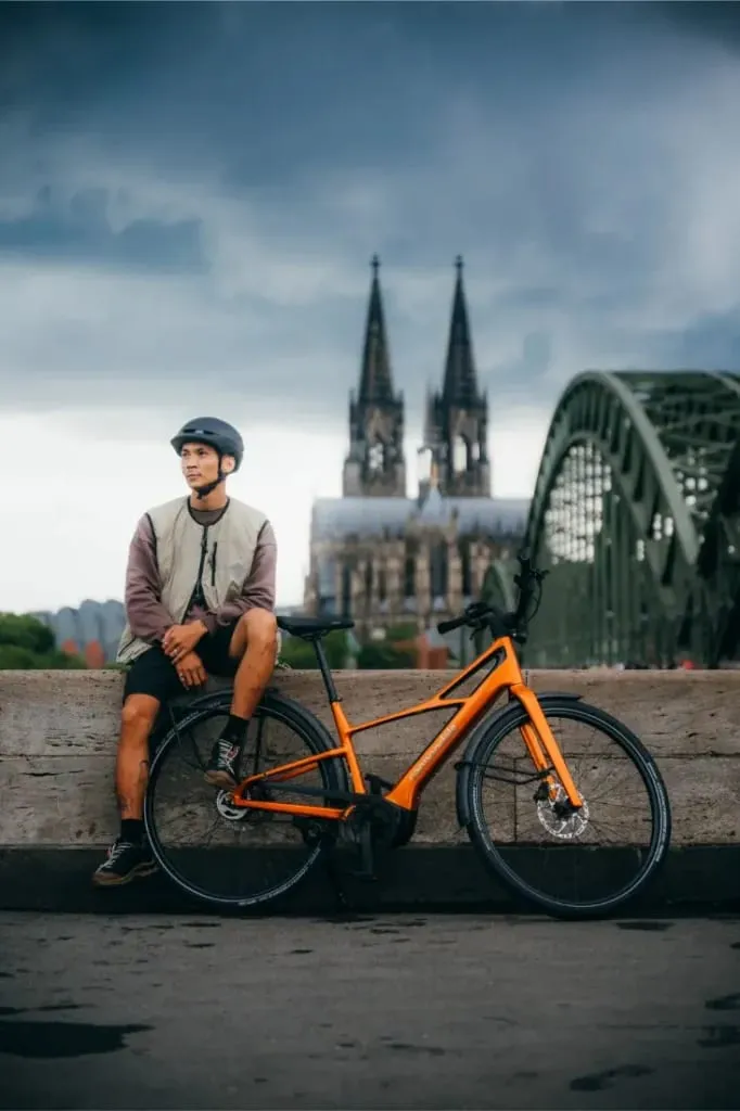 Hombre con casco sentado en un saliente junto a una bicicleta Cannondale FlyingV naranja, con una catedral y un puente al fondo.