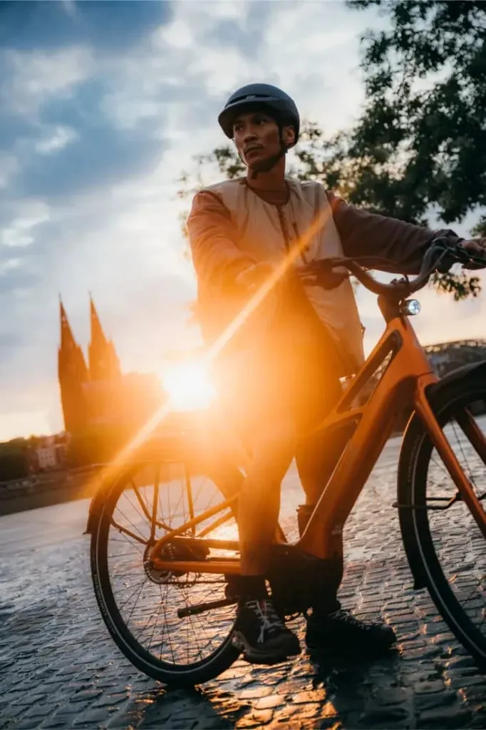 Hombre con casco y bicicleta Cannondale FlyingV al atardecer, paisaje urbano y catedral al fondo, la luz del sol brilla a través de las ruedas.