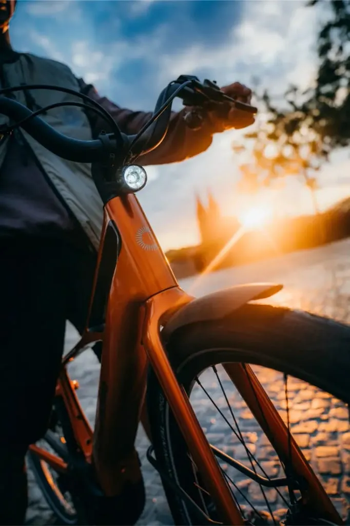 Primer plano de una persona sujetando una bicicleta Cannondale FlyingV naranja al atardecer, con la luz de la bicicleta encendida.
