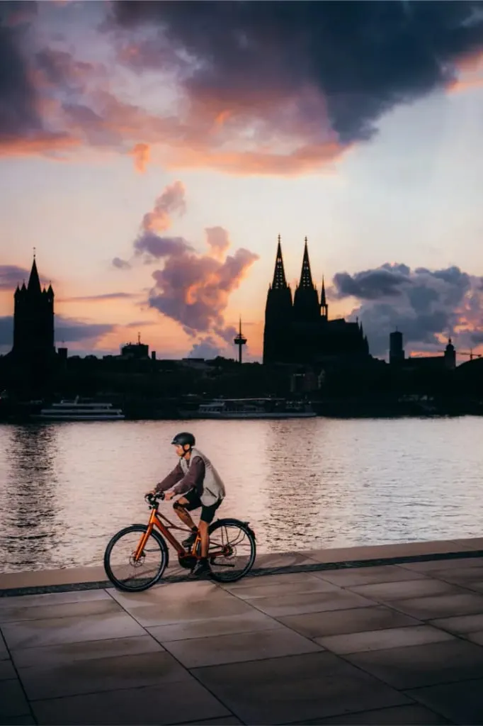 Una persona monta en una bicicleta Cannondale FlyingV junto a un río al atardecer, con el perfil de una ciudad y una catedral de fondo.