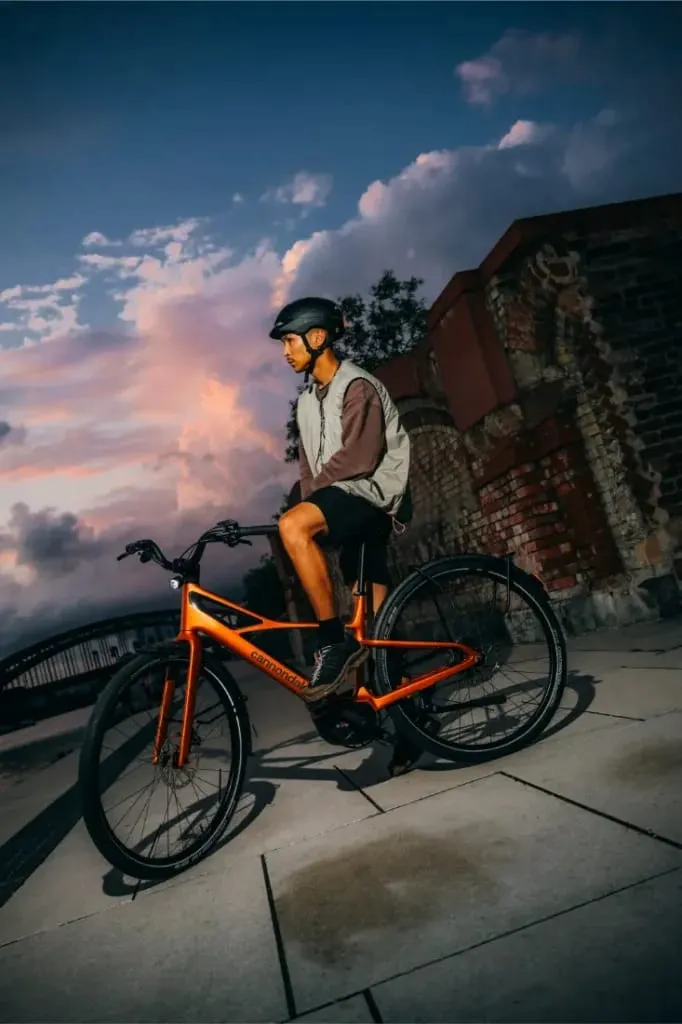 Un hombre con casco monta en una bicicleta Cannondale FlyingV naranja cerca de un muro de ladrillo al atardecer.